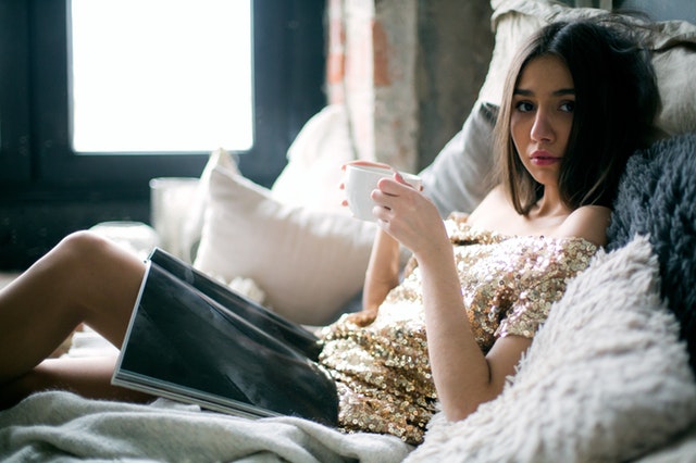 Žena v zlatom lesklom tričku sedí v posteli, číta časopis a pije kávu.jpg